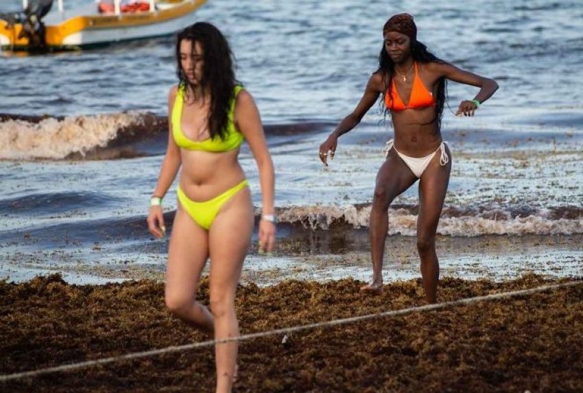 Dos turistas extranjeras regersan del mar y caminan descompuestas entre el sargazo en la playa.